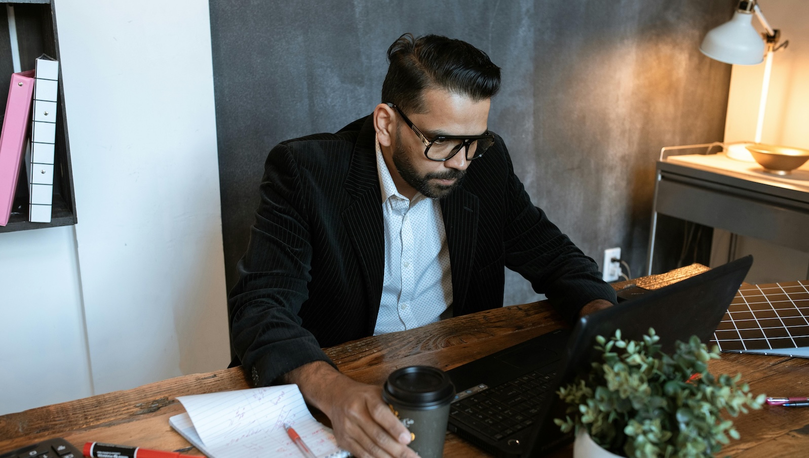 Person with dark hair and in a black suit sitting at a desk with a computer in front of him looking stern